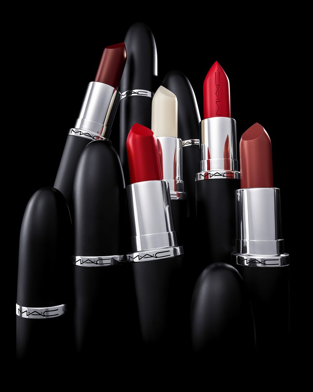 Multiple MAC lipsticks in various shades including red, brown, black, and white are arranged on a black surface, creating a visually appealing makeup still life.
