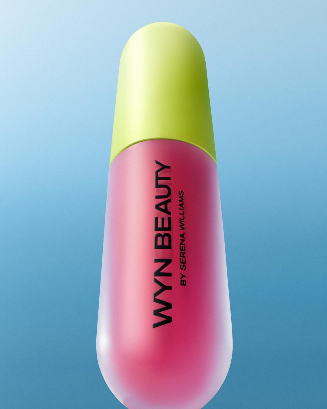 Close-up of a Wyn Beauty lip gloss bottle with a bright green lid and pink base on a blue gradient background. Beauty shot.