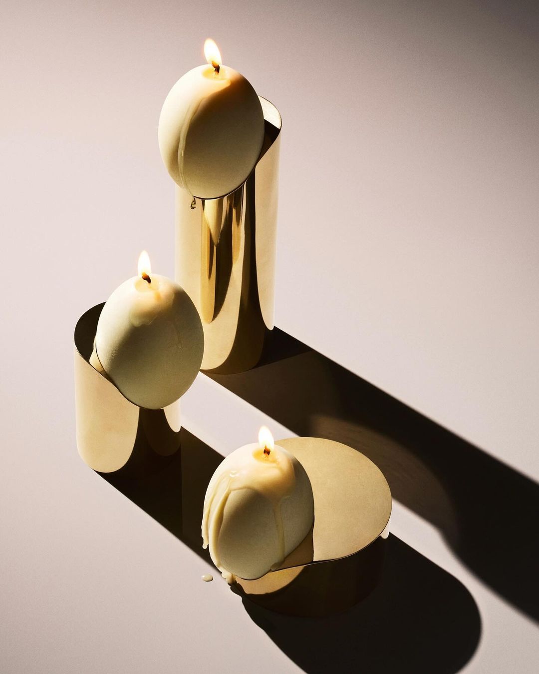 Three lit candles in gold holders on a light surface casting a shadow, ideal for product photography or home decor concepts.