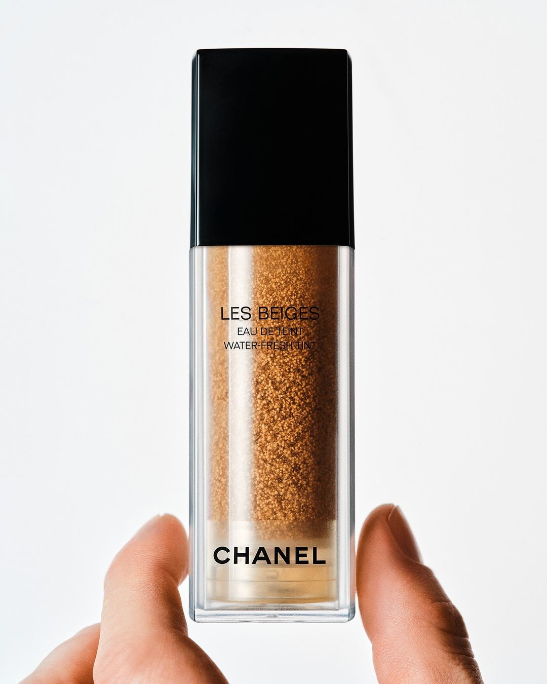 Tan tinted makeup in a clear bottle with a black lid held by a hand, against a white background, product shot for the beauty industry.