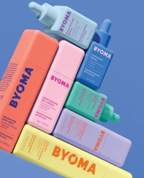 Colorful Byoma skincare bottles and tubes stacked diagonally against a solid blue background by Rikky Fernandes