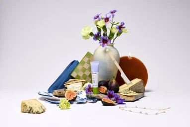Still life arrangement of a skincare tube, figs, cheese, crackers, flowers in a ceramic vase, and a burning candle on a white surface.