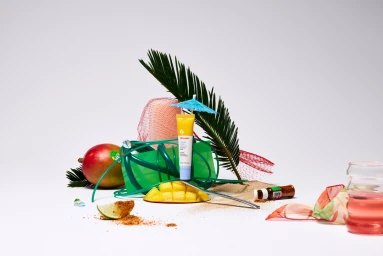 A yellow cosmetics tube stands upright on a mango slice amid tropical props and sand against a white studio background.