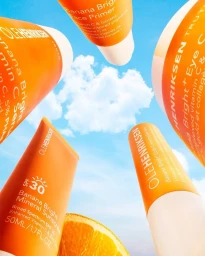 Orange skincare bottles and a fresh orange slice angled upward against a bright blue sky with white clouds.