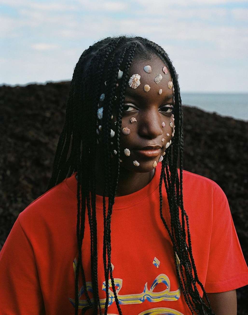 Portrait of a young woman with seashell face decorations, braided hair, and a red t-shirt, posing outdoors against a nature background.