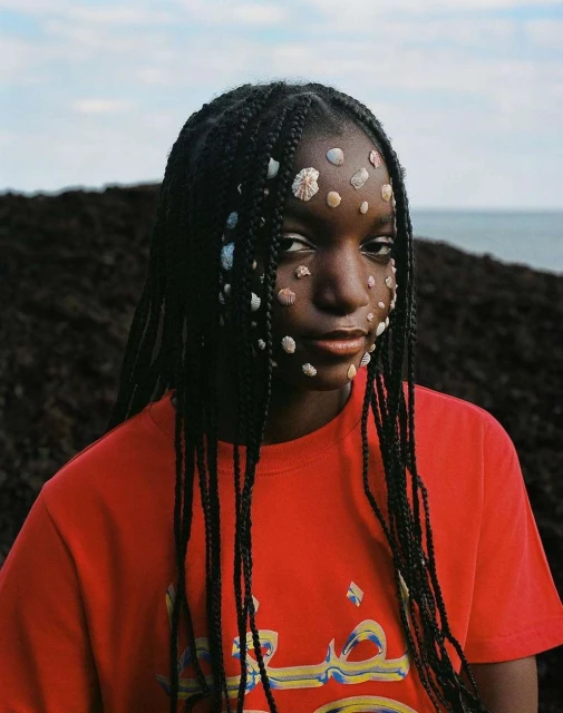 Portrait of a young woman with seashell face decorations, braided hair, and a red t-shirt, posing outdoors against a nature background.