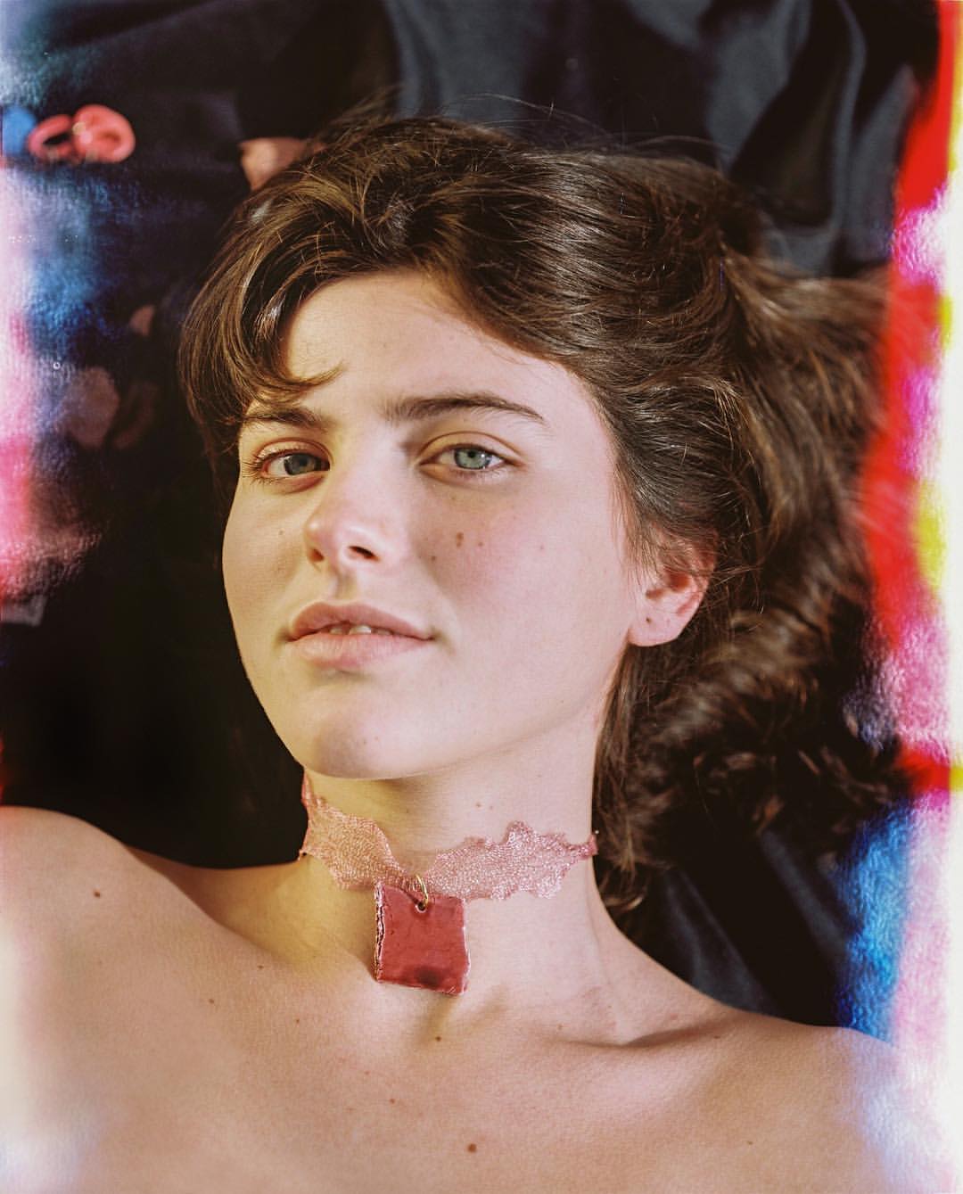 Close-up portrait of a fair-skinned young woman with heterochromia wearing a lace choker necklace against a dark, colorful background.