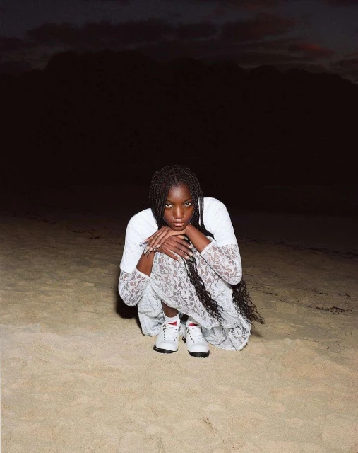 A young woman with long braids poses outdoors on a sandy surface, wearing a white top and lacy dress, with a dark, cloudy sky above.