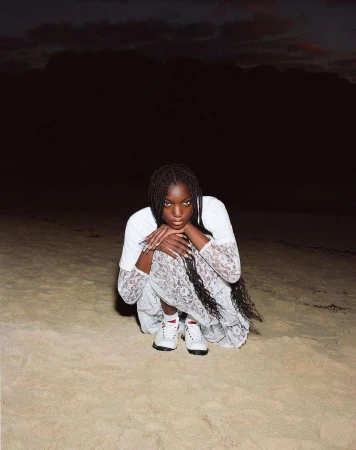 A young woman with long braids poses outdoors on a sandy surface, wearing a white top and lacy dress, with a dark, cloudy sky above.