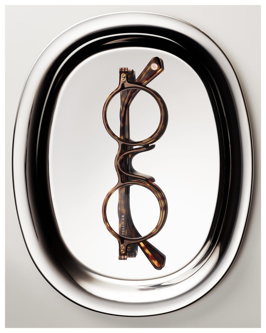 Optical eyeglasses with tortoise shell frames are displayed on an oval silver tray against a light backdrop in a studio shot.