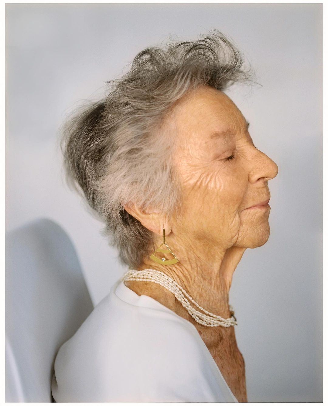 Profile portrait of an elderly woman with gray hair, wearing a white top and multi-strand pearl necklace, and triangular gold earrings.