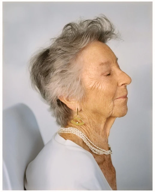 Profile portrait of an elderly woman with gray hair, wearing a white top and multi-strand pearl necklace, and triangular gold earrings.
