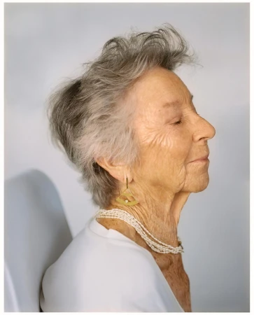 Profile portrait of an elderly woman with gray hair, wearing a white top and multi-strand pearl necklace, and triangular gold earrings.