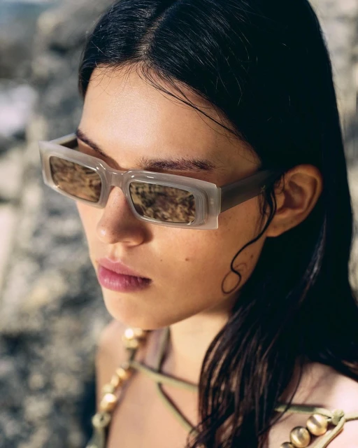 Close-up of an adult woman with dark hair, wearing sunglasses and a beaded necklace, posing against a neutral stone background outdoors.