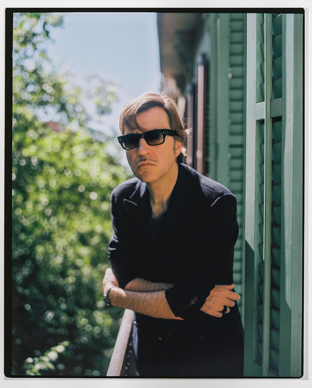 Adult male wearing sunglasses stands on a balcony with green shutters, wearing a black coat, and looking at the camera with a neutral expression.