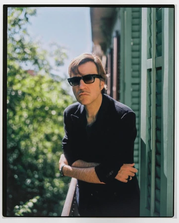 Adult male wearing sunglasses stands on a balcony with green shutters, wearing a black coat, and looking at the camera with a neutral expression.