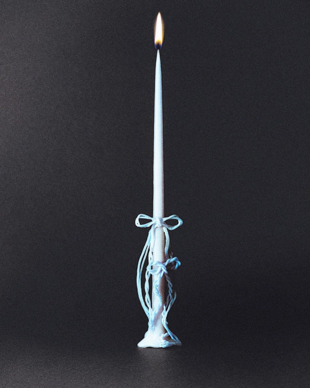 Lit white candle on a black background with a decorative light-blue bow and dripping wax at the base.