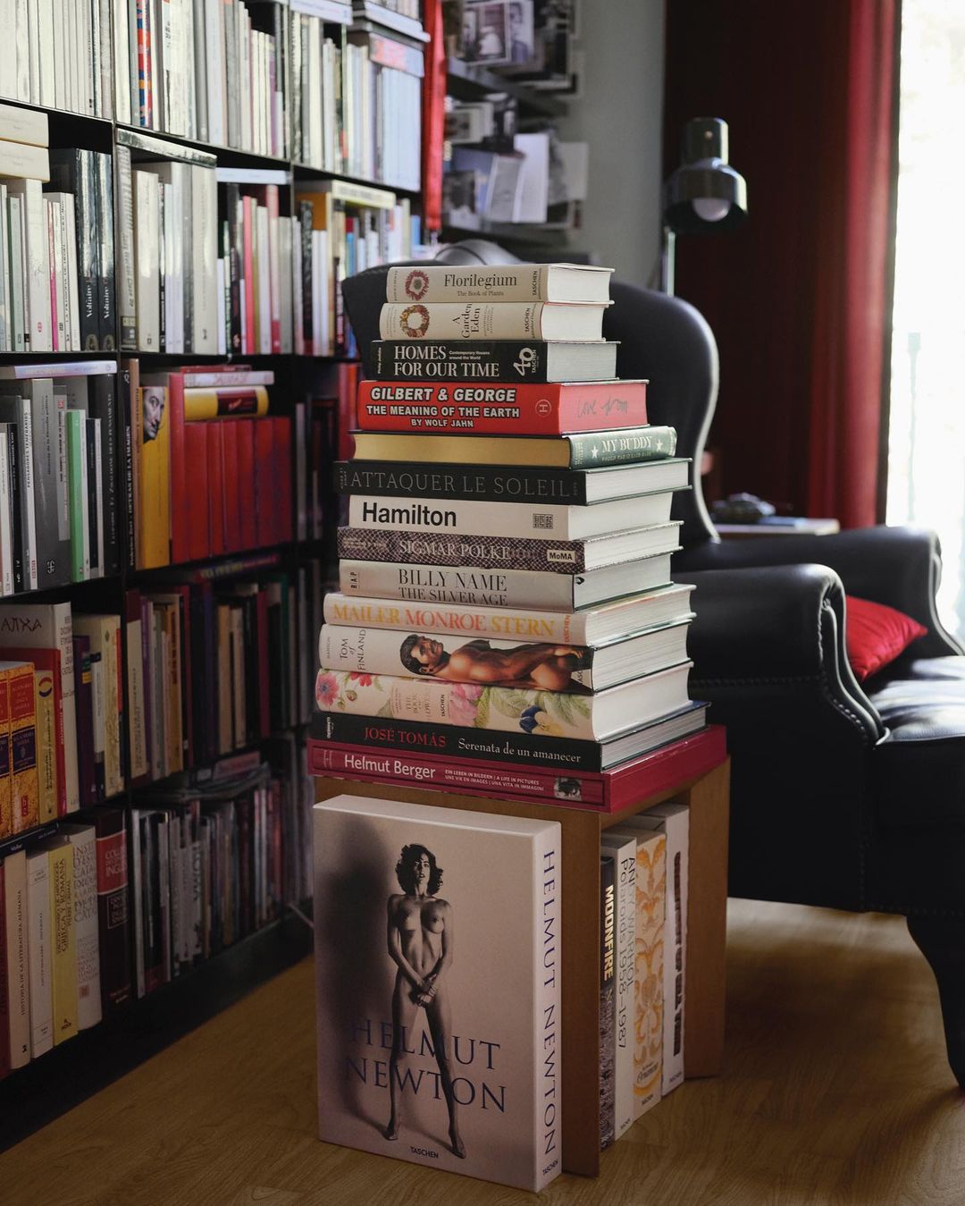 A stack of assorted books on a wooden stool, with a large bookshelf visible in the background. An armchair is set to the side.