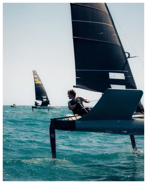 A sailboat race featuring a man steering a black sailboat on a wavy ocean under a sunny sky with another boat in the background.