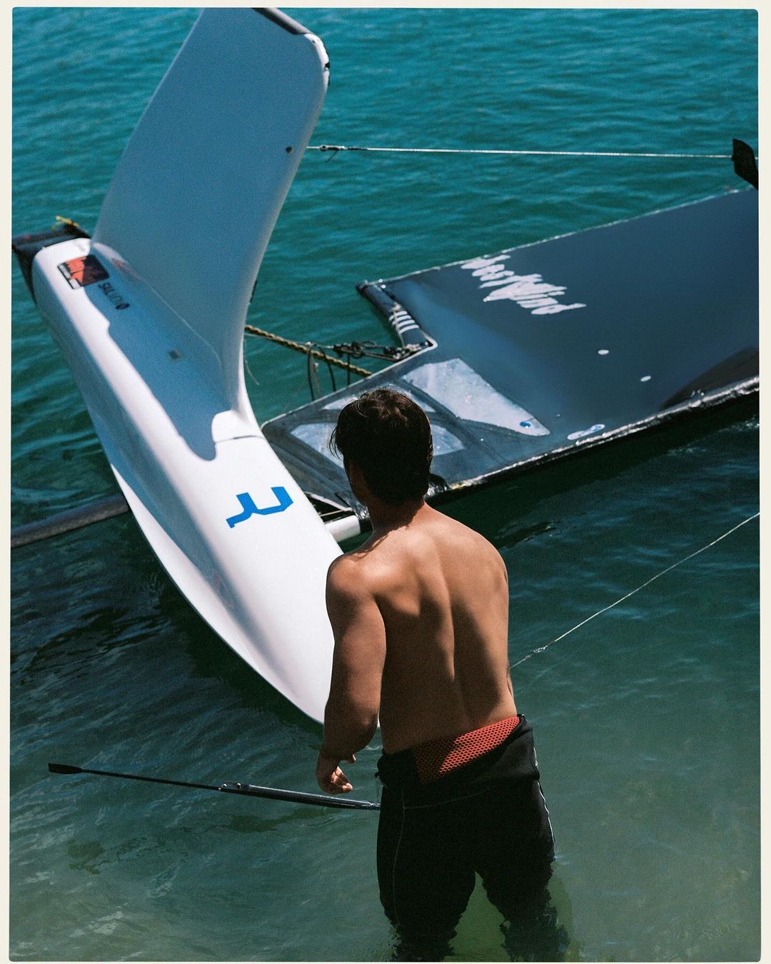 A shirtless adult male stands in turquoise water next to a white catamaran sailboat. Sunny, bright daylight. Water sport theme.
