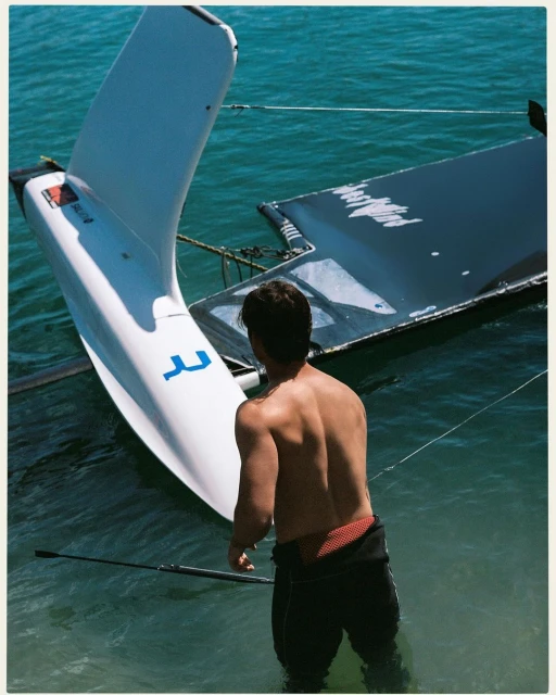 A shirtless adult male stands in turquoise water next to a white catamaran sailboat. Sunny, bright daylight. Water sport theme.