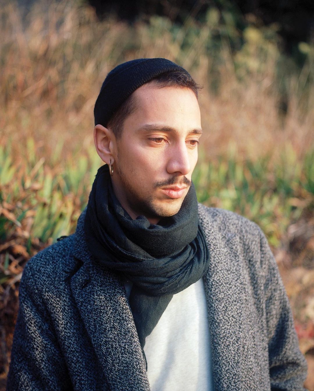A portrait of a man wearing a beanie and scarf, looking down, with a coat, set against a background of tall grasses and soft natural light.