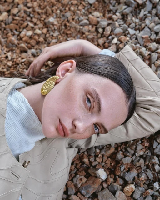 Woman with slicked-back hair and large gold earring lies among brown rocks wearing a tan vest and a white top in a portrait-style shot.