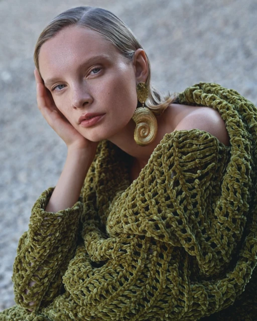 Close-up portrait of a blonde woman with large spiral earrings draped in a green, woven off-the-shoulder garment, outdoors.