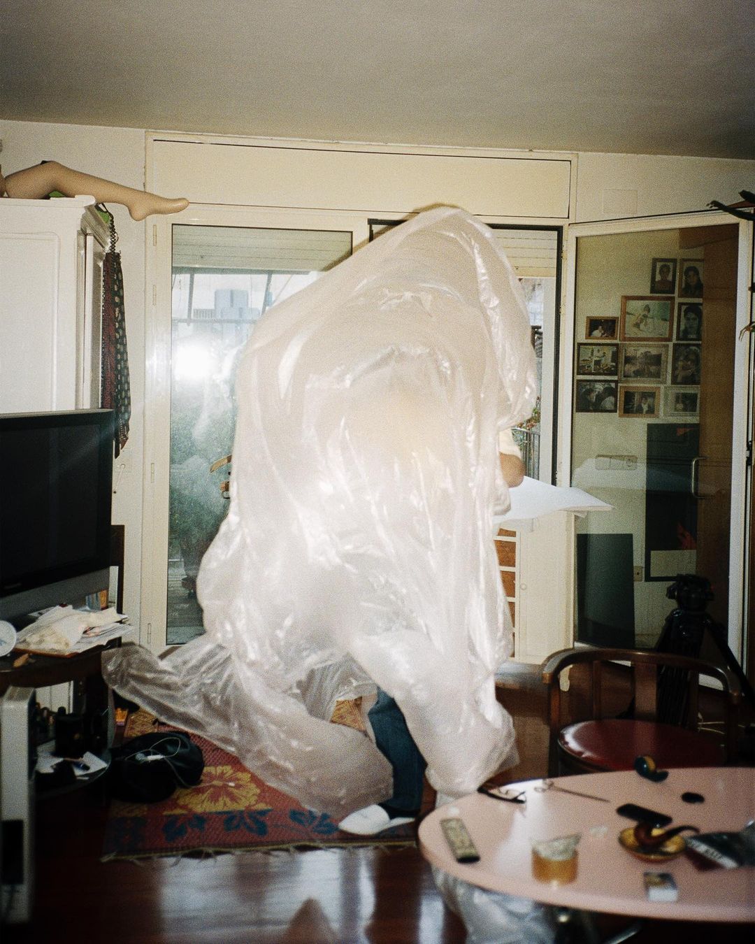 Person covered in translucent plastic material stands in an indoor setting, surrounded by furniture and household objects, creating an intriguing scene.
