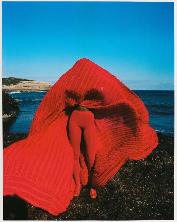 Model standing on a dark rocky coastline under a clear blue sky holding a large billowing red textured fabric garment
