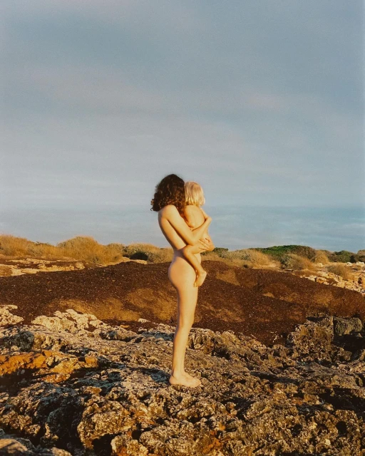 A nude adult female stands on a rocky landscape holding a small child against a blue sky, creating a striking and intimate scene.