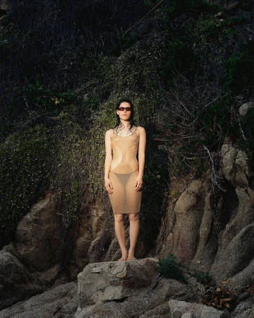 Woman in sheer nude slip dress standing barefoot on a rocky coastal cliffside in Barcelona