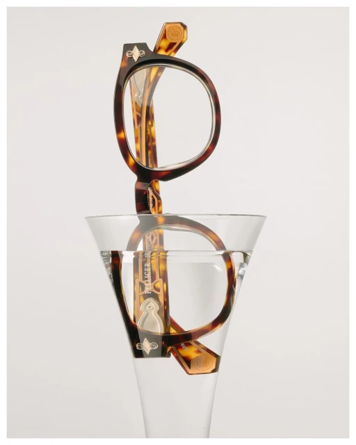 Tortoiseshell eyeglasses are partially submerged in a martini glass filled with water, against a plain white background, in a product shot.