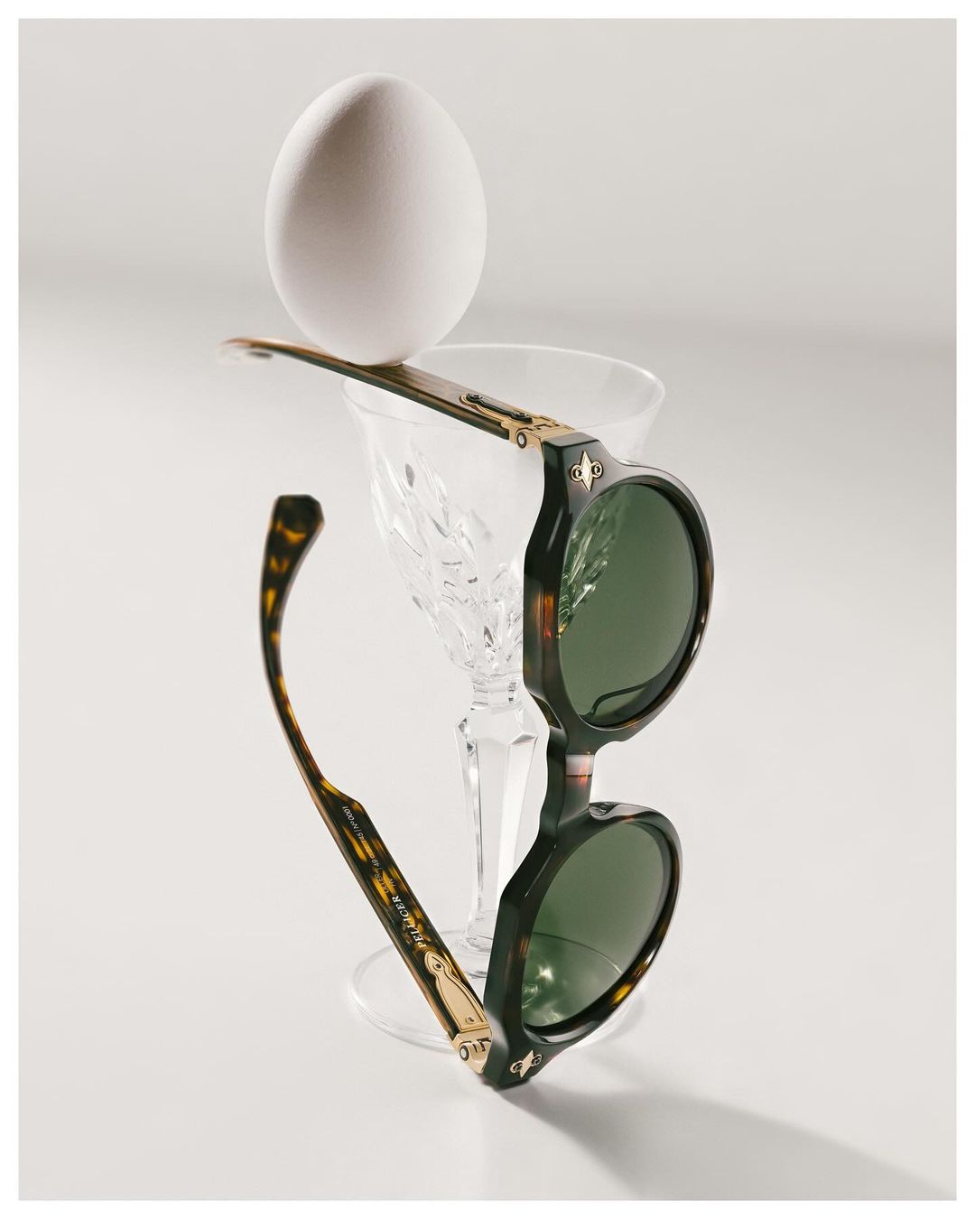 Round sunglasses with tortoise frames balance a white egg atop a crystal glass in a bright, minimalist composition on a white surface.
