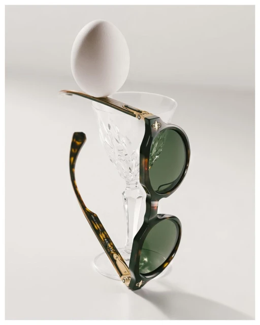 Round sunglasses with tortoise frames balance a white egg atop a crystal glass in a bright, minimalist composition on a white surface.
