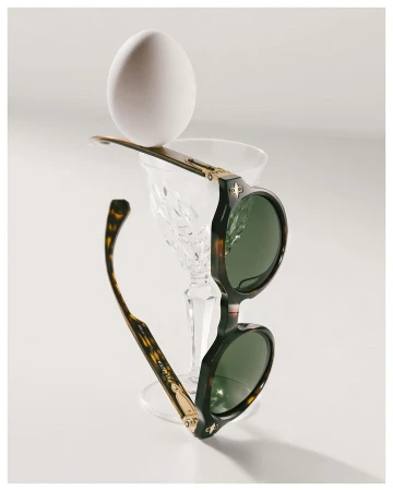 Round sunglasses with tortoise frames balance a white egg atop a crystal glass in a bright, minimalist composition on a white surface.