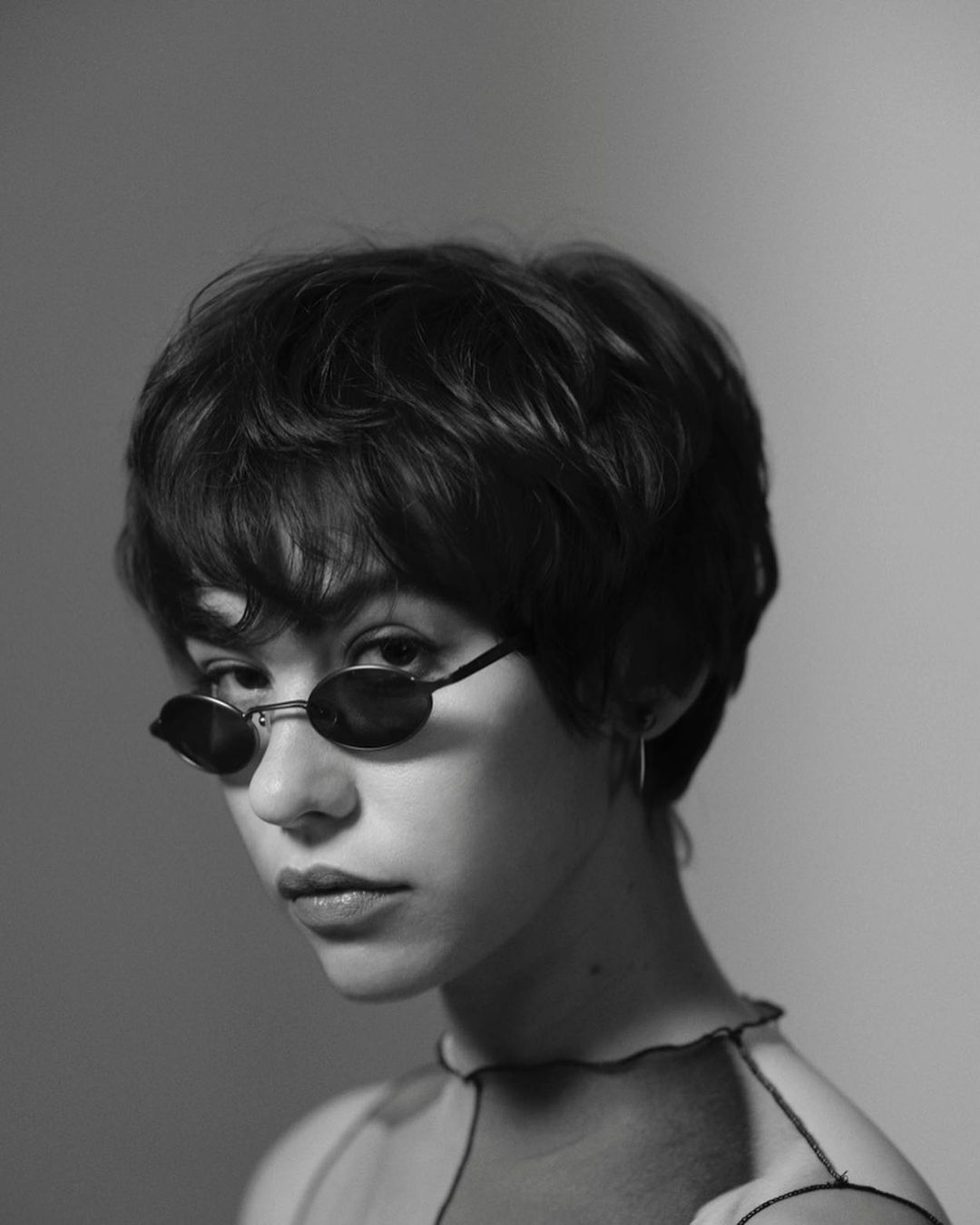 Black and white portrait of a young adult woman with a short haircut and sunglasses, looking at the camera in an indoor setting.