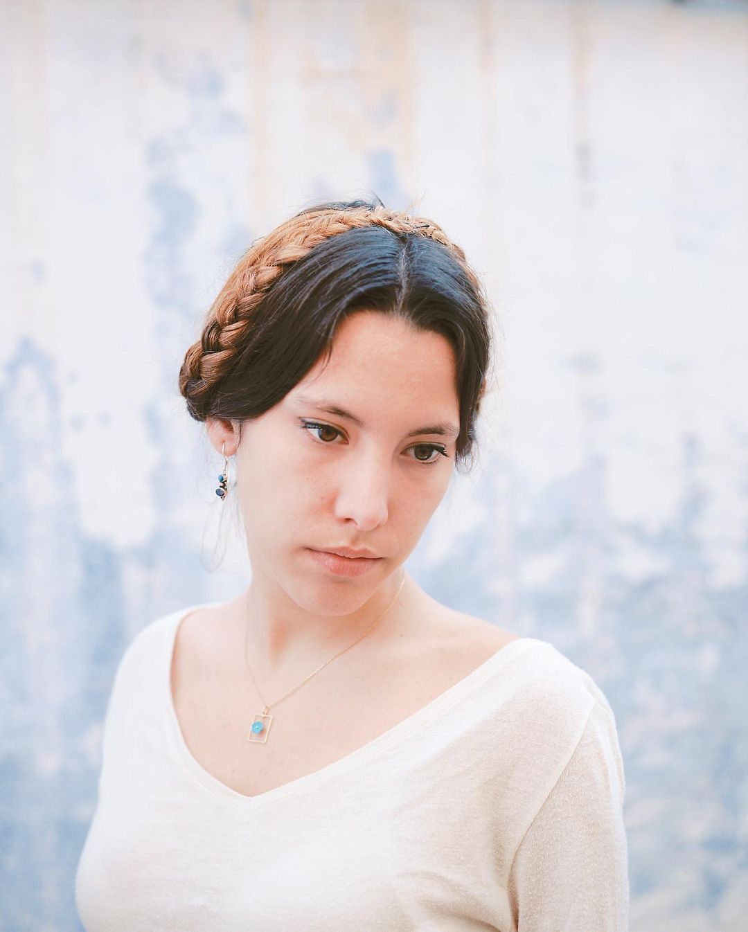 Portrait of a woman with brown braided hair wearing a V-neck shirt, necklace, and earrings, posing in front of a distressed blue wall.