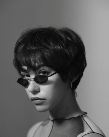 Close-up black and white portrait of a young woman wearing dark oval sunglasses and a sheer top with exposed seams by Rita Puig-serra.
