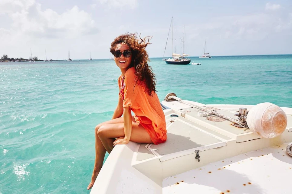 A woman with curly hair sits on a boat in the ocean, wearing sunglasses and an orange cover-up, with sailboats visible in the background.