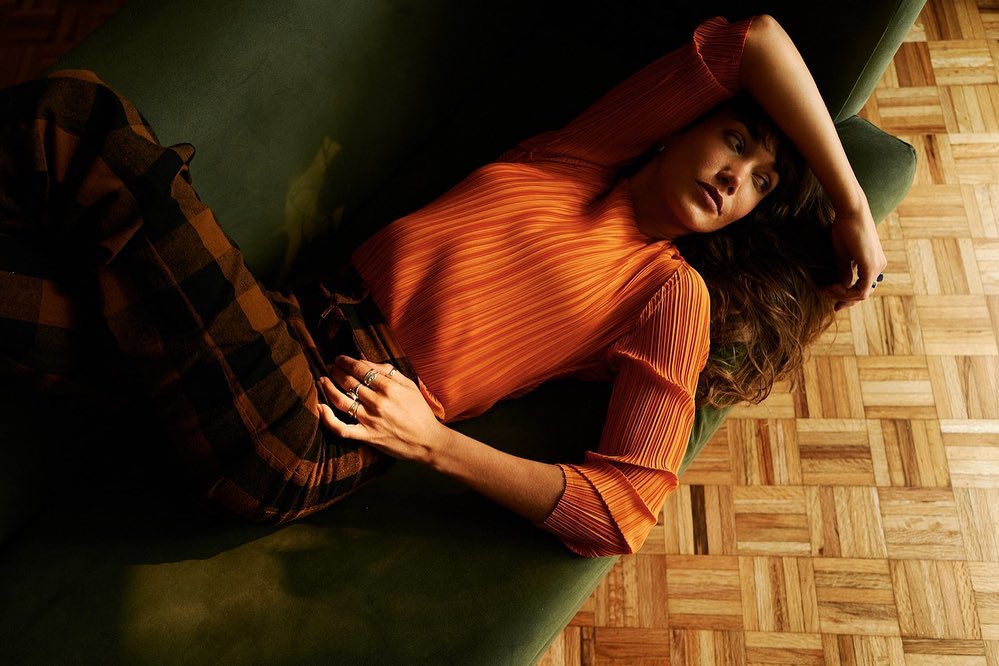 Brunette woman in an orange blouse and plaid pants lies on a green couch on a parquet floor in a lifestyle fashion portrait.