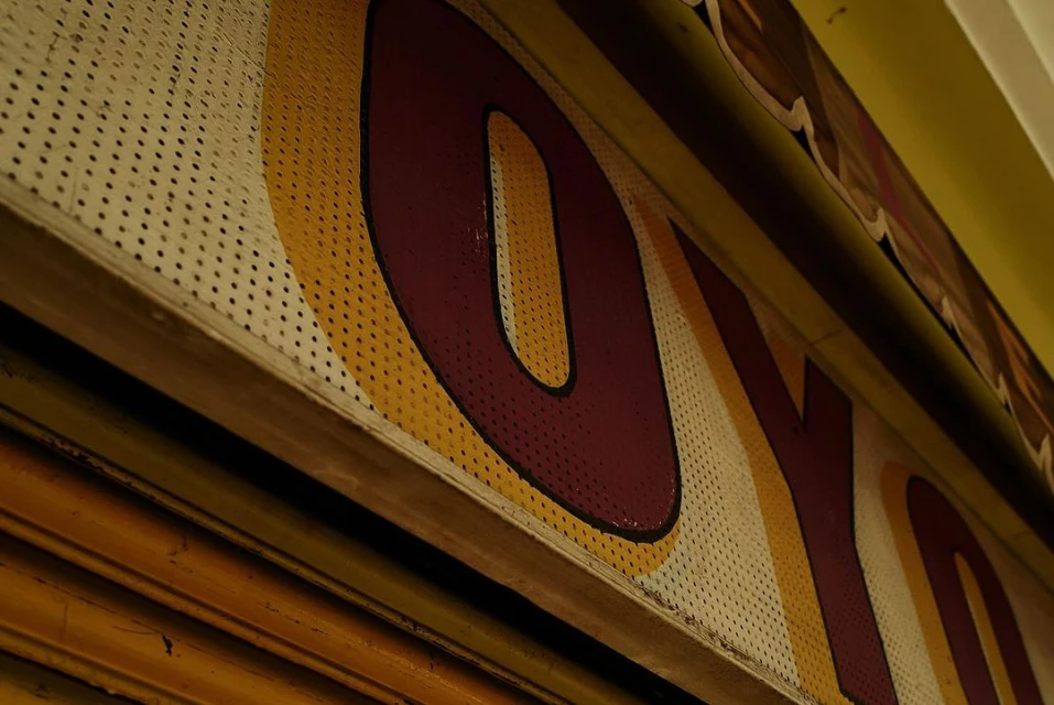 Close-up of weathered sign with maroon letters and yellow stripes on perforated white background, wooden frame along yellow wall.