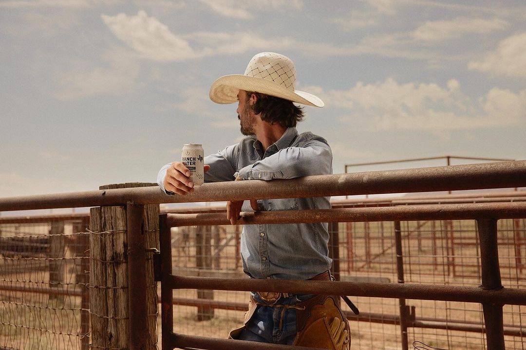 Man in cowboy hat leans against fence, holding a Ranch Water can, wearing denim shirt and chaps, with a tan desert landscape background.
