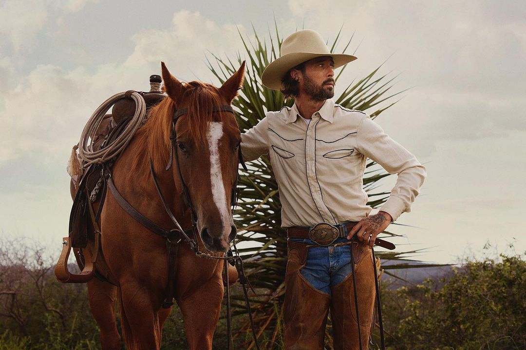 A man in a cowboy hat stands next to a brown horse in an outdoor setting, wearing a Western-style shirt and chaps, under an overcast sky.