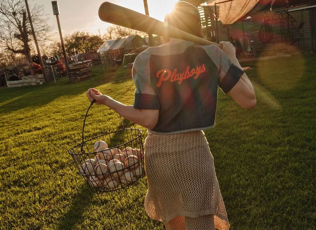 A woman wearing a baseball-style crop top and skirt carries a wire basket full of baseballs, with a bat over her shoulder in a grassy outdoor setting.
