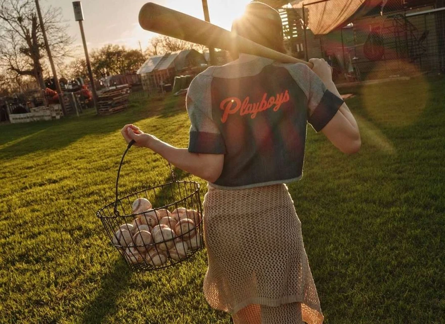 A woman wearing a baseball-style crop top and skirt carries a wire basket full of baseballs, with a bat over her shoulder in a grassy outdoor setting.