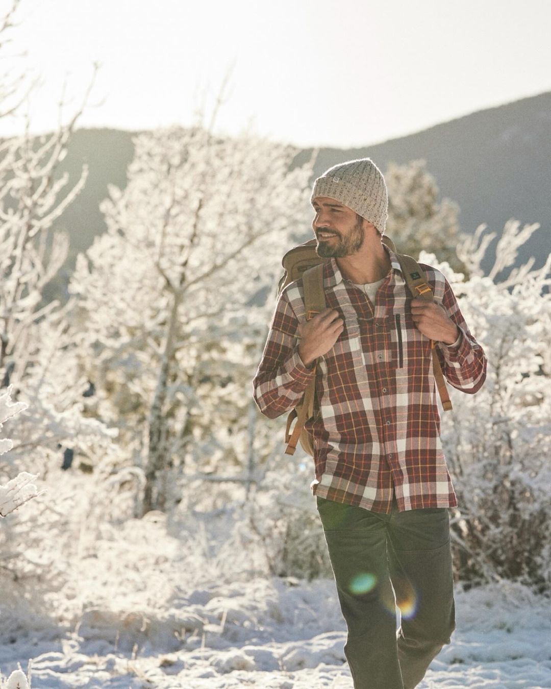 Man with backpack hikes in snowy outdoors wearing a plaid shirt, knit hat and dark pants on a sunny winter day.