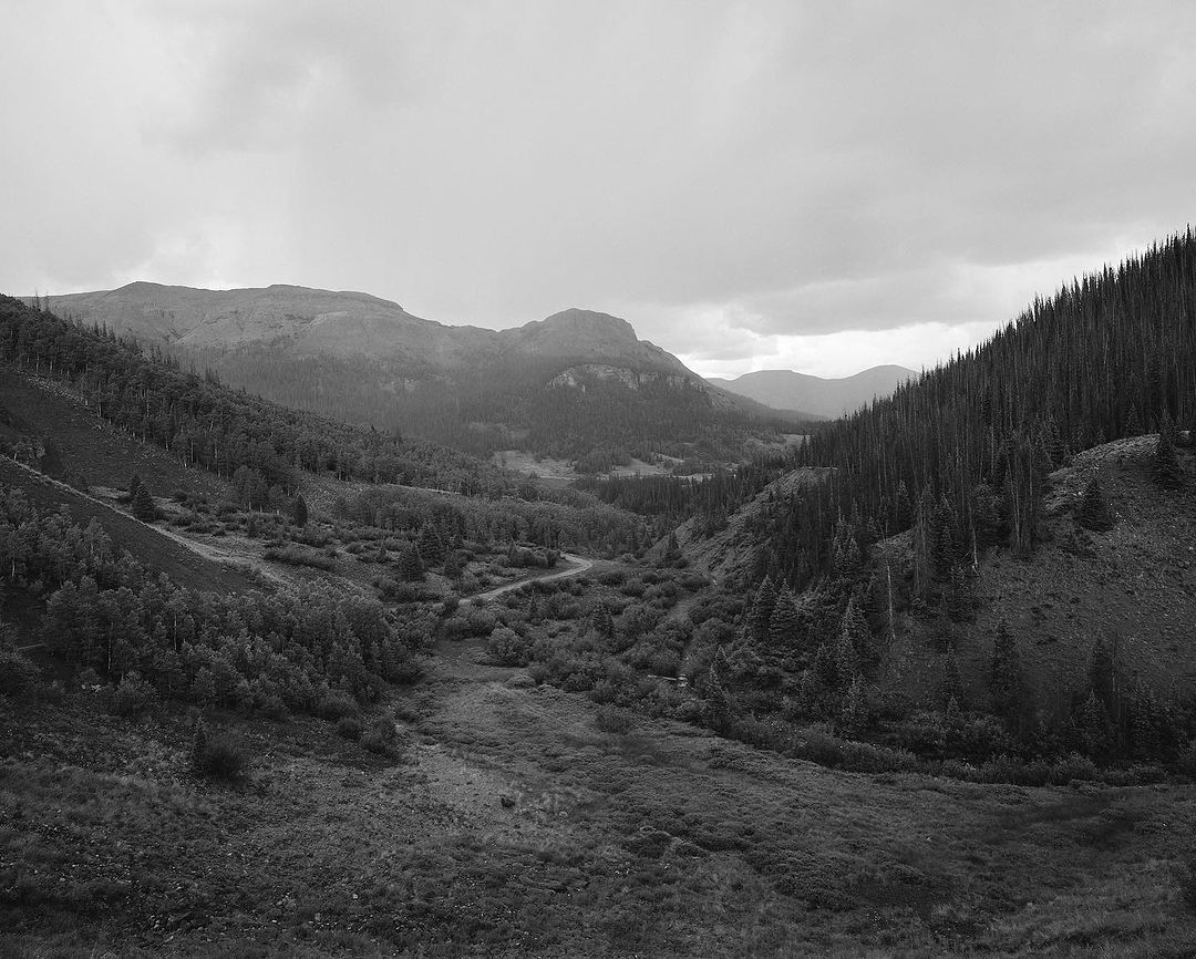 Black and white landscape featuring a winding road through a valley, surrounded by mountains, dense forests, and a cloudy sky overhead.