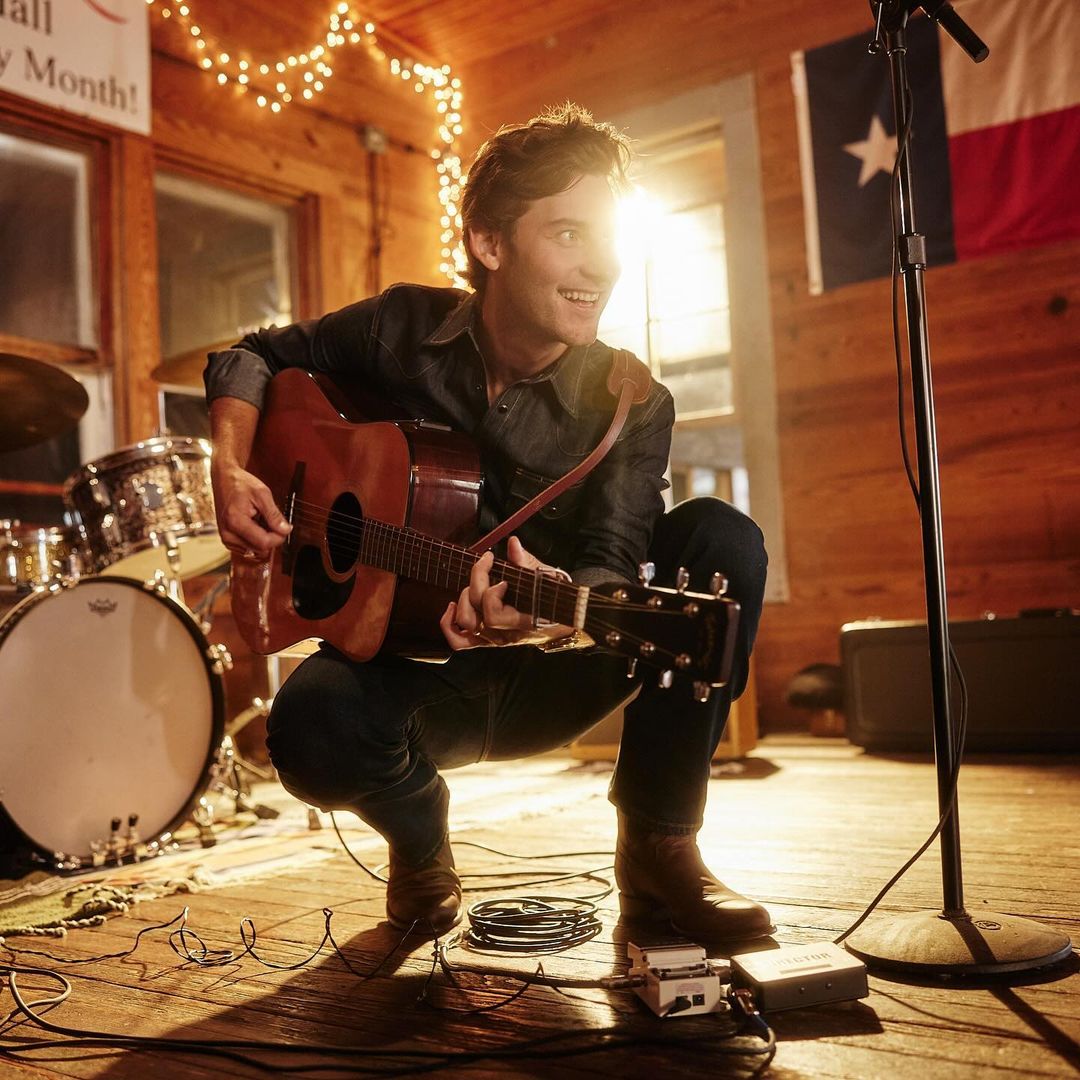 Man plays acoustic guitar on stage with drum set and Texas flag in the background, bathed in warm, vibrant lighting.