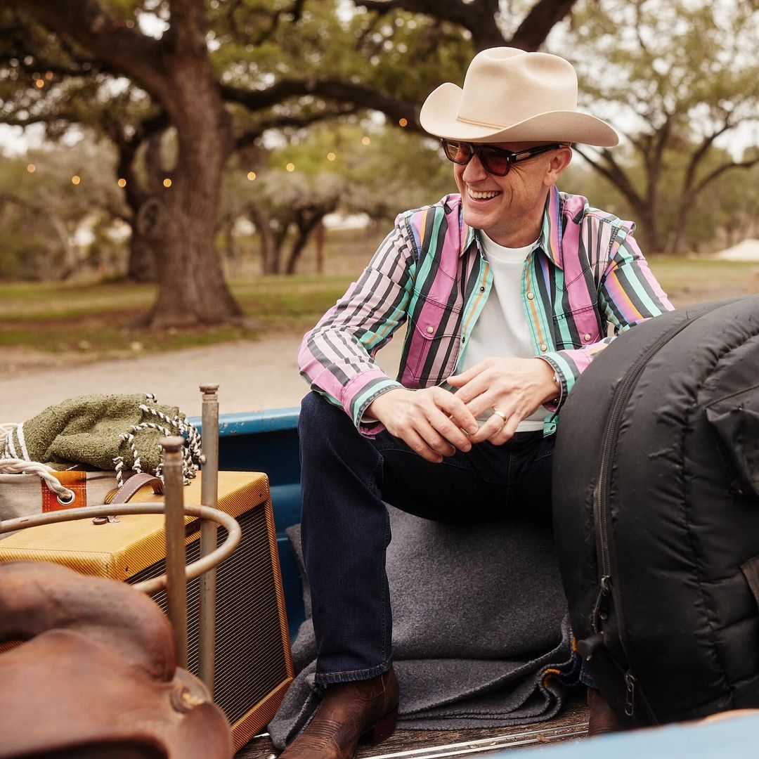 Smiling man in cowboy hat and striped shirt sits in the back of a truck with luggage and musical equipment, outdoor setting.
